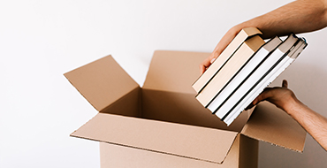 A hand placing a stack of books inside a brown cardboard box.